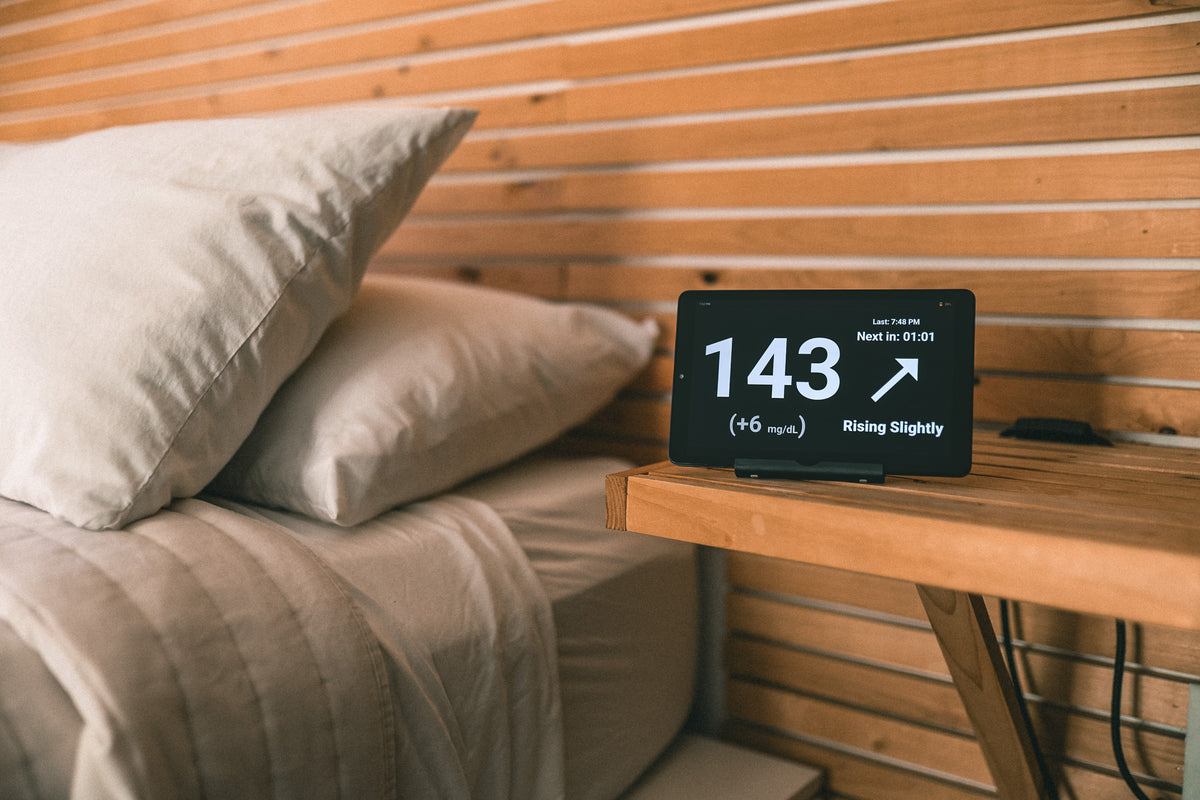 Close-up of the display on a wooden shelf: the large-number view in dark mode, a white 143 with a rising arrow, plus 6 milligrams per decilitre, and the time of the last reading.